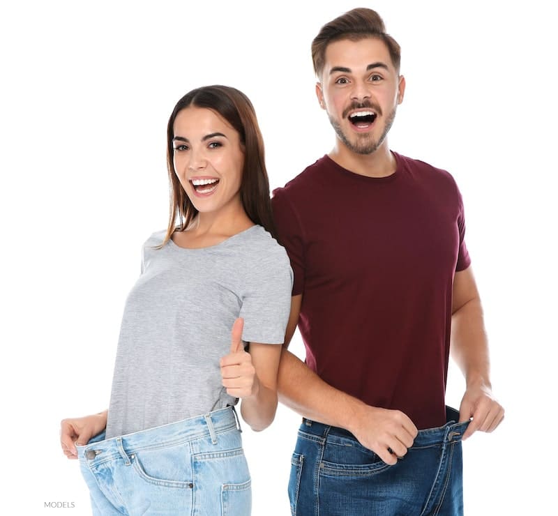 Young and attractive couple smiling and laughing as they hold out large pants after weight loss.