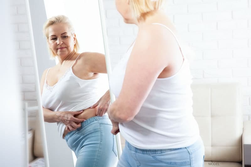 Middle-aged woman looking in a mirror and pinching excess fat and skin in her midsection